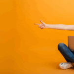 Cheerful woman in casual clothing and eyeglasses sitting with a cardboard box filled with Halloween decorations, smiling at the camera. Illustrates festive preparation for seasonal email campaigns.