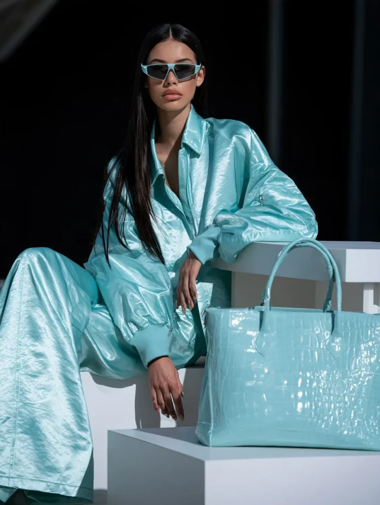 Woman in a pale aqua satin bomber jacket and wide-leg trousers sitting beside a matching crocodile-embossed tote bag